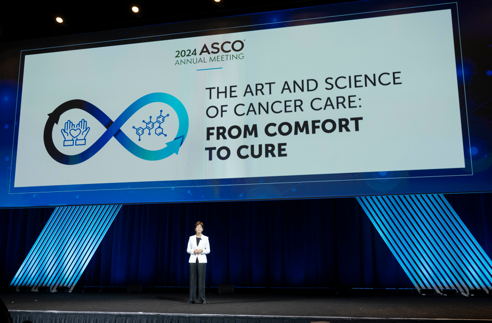 Lynn Schuchter stands on stage at the ASCO Annual Meeting with a large screen displaying the theme, "The Art and Science of Cancer Care: From Comfort to Cure."